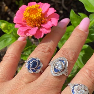 May include: Three silver-toned rings featuring blue and clear stones. The first ring has a central oval blue stone surrounded by smaller blue stones. The second ring features a central round clear stone with blue accents. The third ring has a round clear stone with blue detailing.