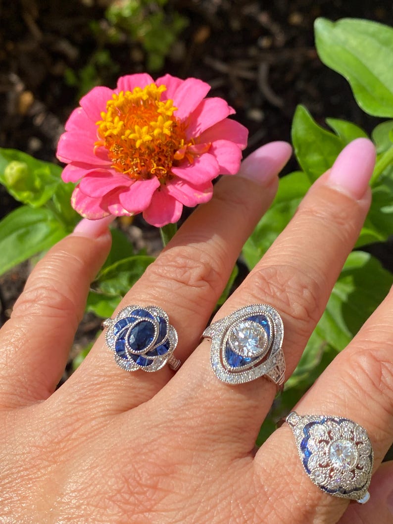 May include: Three silver-toned rings featuring blue and clear stones. The first ring has a central oval blue stone surrounded by smaller blue stones. The second ring features a central round clear stone with blue accents. The third ring has a round clear stone with blue detailing.