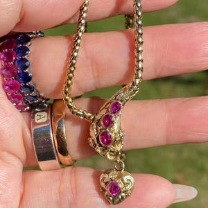 May include: A gold-toned necklace with a pendant featuring a heart-shaped design and embedded pink gemstones. The necklace is held up, showcasing its intricate details. A ring with the letter "A" is also visible.