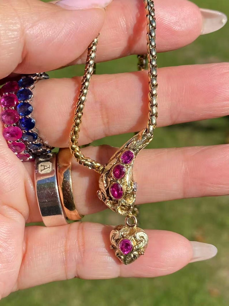 May include: A gold-toned necklace with a pendant featuring a heart-shaped design and embedded pink gemstones. The necklace is held up, showcasing its intricate details. A ring with the letter "A" is also visible.