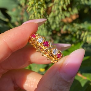 May include: A gold ring featuring alternating red and clear gemstones. The ring has a woven design with the gemstones set in the center. The ring is held up in front of a green and yellow background.