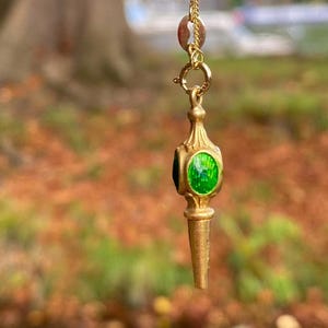 Könnte beinhalten: Goldfarbener Anhänger mit einem leuchtend grünen, ovalen Mittelstein. Der Anhänger hängt an einer feinen Goldkette. Der unscharfe Hintergrund zeigt grüne und braune Farbtöne.