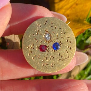 May include: A circular, gold-coloured brooch with detailed floral patterns. It has three inset gemstones: a clear, a red, and a blue stone. The brooch is held in a hand, with a blurred background of leaves and grass.