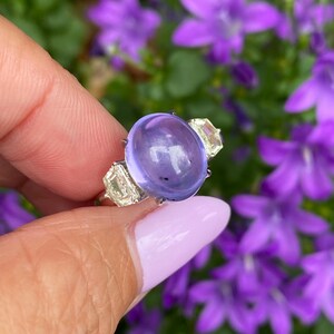 May include: A silver ring featuring a large, oval, translucent, purple gemstone as the centerpiece. Two smaller, rectangular, clear gemstones flank the main stone. The ring is held against a backdrop of purple flowers.