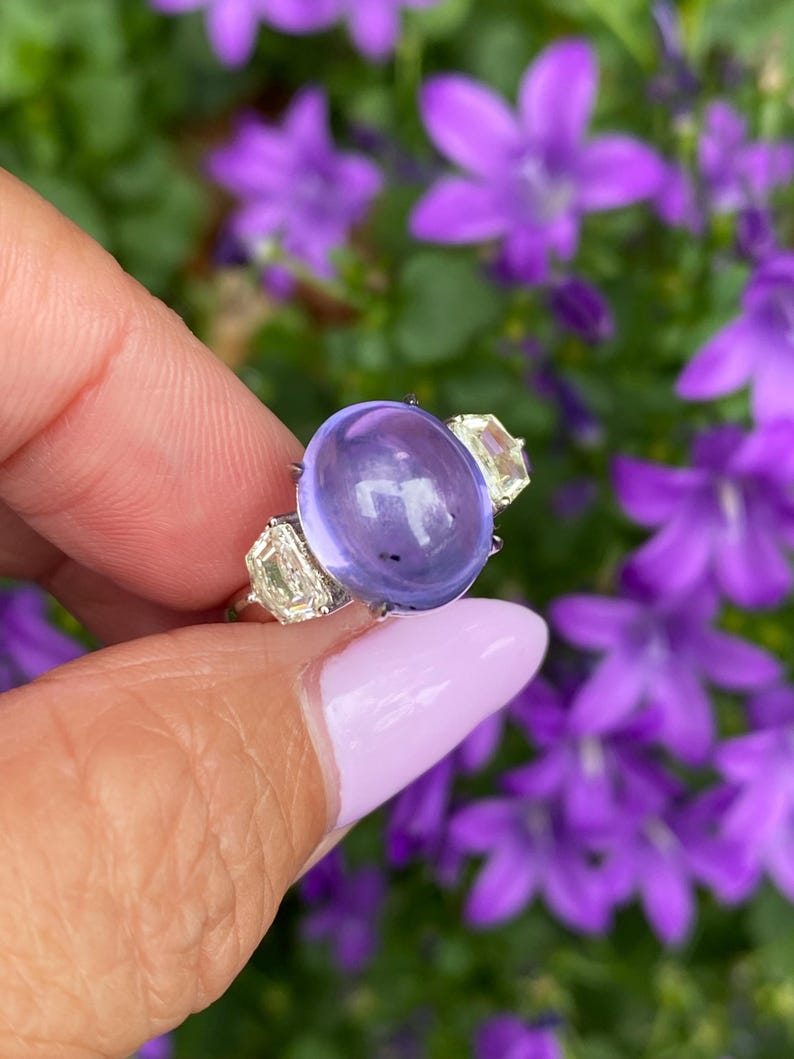 May include: A silver ring featuring a large, oval, translucent, purple gemstone as the centerpiece. Two smaller, rectangular, clear gemstones flank the main stone. The ring is held against a backdrop of purple flowers.