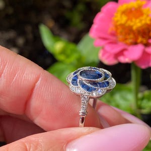 May include: A silver-toned ring featuring a central oval blue sapphire surrounded by a floral-shaped halo of smaller blue sapphire baguettes and tiny clear accent stones, set in a metal band with intricate milgrain detailing along the edges.