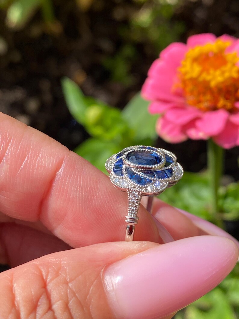 May include: A silver-toned ring featuring a central oval blue sapphire surrounded by a floral-shaped halo of smaller blue sapphire baguettes and tiny clear accent stones, set in a metal band with intricate milgrain detailing along the edges.