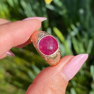 以下が含まれることがあります： 花柄が刻まれたゴールドのリング。大きなファセットカットのルビーがベゼルセッティングで留められています。