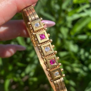 Può includere: Un bracciale rigido a cerniera in oro caratterizzato da una fila di cinque castoni quadrati. I castoni alternano pietre bianche trasparenti e pietre rosa vivaci, arricchiti da piccole sfere d'oro e dettagli in filo metallico.