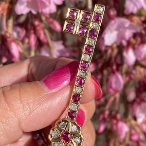 May include: A gold key-shaped pendant with a ruby and diamond design. The key has a flower-shaped end with a large ruby in the center. The key is surrounded by pink flowers.