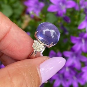 May include: A silver ring featuring a large, round, translucent, purple gemstone. Two smaller, triangular, clear gemstones flank the main stone. The ring is held against a backdrop of purple flowers.