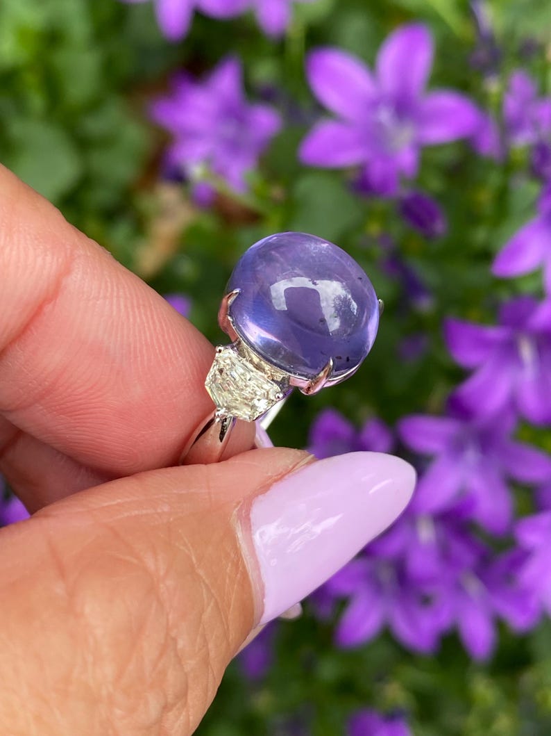 May include: A silver ring featuring a large, round, translucent, purple gemstone. Two smaller, triangular, clear gemstones flank the main stone. The ring is held against a backdrop of purple flowers.