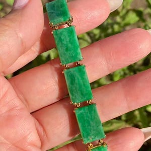 Könnte beinhalten: Ein Jade-Armband mit rechteckigen grünen Jade-Steinen, die durch goldfarbene Glieder verbunden sind. Der polierte Jade hat eine lebendige grüne Farbe. Das Armband wird vor einem natürlichen Hintergrund präsentiert.
