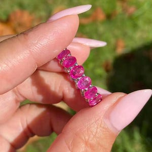 May include: A silver ring set with a line of oval-cut, bright pink gemstones. The ring is held up against a blurred green and brown background, highlighting the vivid colour of the stones.