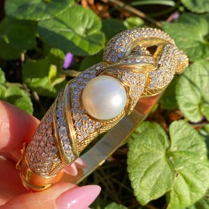 May include: A gold-toned bracelet with a large, round pearl and encrusted with small, sparkling stones. The bracelet has a knot design at the top and is held against a background of green foliage.