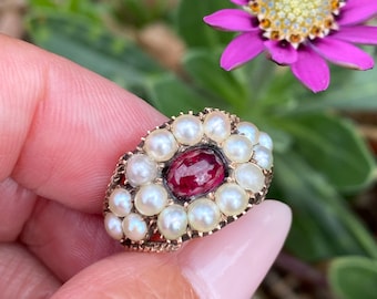May include: A gold ring featuring a central oval, deep red gemstone surrounded by small, white pearls. The ring is held in a hand, with a blurred background of green foliage and a vibrant purple flower.
