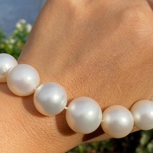 Pode incluir: Um close-up de um colar de p&eacute;rolas brancas. As p&eacute;rolas s&atilde;o grandes, redondas e t&ecirc;m um brilho lustroso. O colar &eacute; enfiado em um cord&atilde;o branco. Uma pulseira de prata com uma pedra preta &eacute; vis&iacute;vel no fundo.