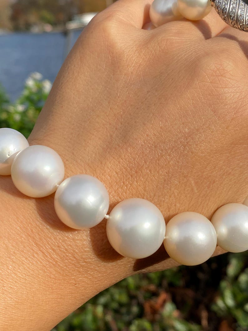 Pode incluir: Um close-up de um colar de p&eacute;rolas brancas. As p&eacute;rolas s&atilde;o grandes, redondas e t&ecirc;m um brilho lustroso. O colar &eacute; enfiado em um cord&atilde;o branco. Uma pulseira de prata com uma pedra preta &eacute; vis&iacute;vel no fundo.