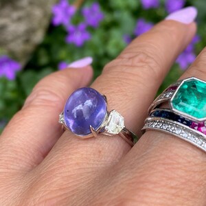 May include: A silver ring featuring a large, oval, purplish-blue gemstone flanked by two smaller, clear, rectangular stones. Another ring with a large, emerald-green, octagonal stone is also visible, set with smaller stones.