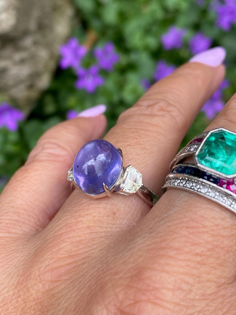 May include: A silver ring featuring a large, oval, purplish-blue gemstone flanked by two smaller, clear, rectangular stones. Another ring with a large, emerald-green, octagonal stone is also visible, set with smaller stones.