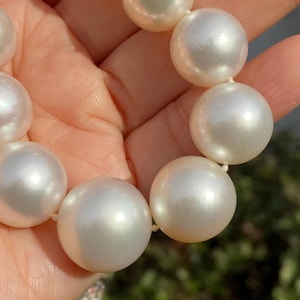 Pode incluir: Um colar de p&eacute;rolas brancas grandes em close-up. As p&eacute;rolas, de forma esf&eacute;rica, s&atilde;o de tamanho uniforme e t&ecirc;m um brilho perolado. Est&atilde;o enfiadas em um fio fino. O fundo desfocado sugere um ambiente externo.
