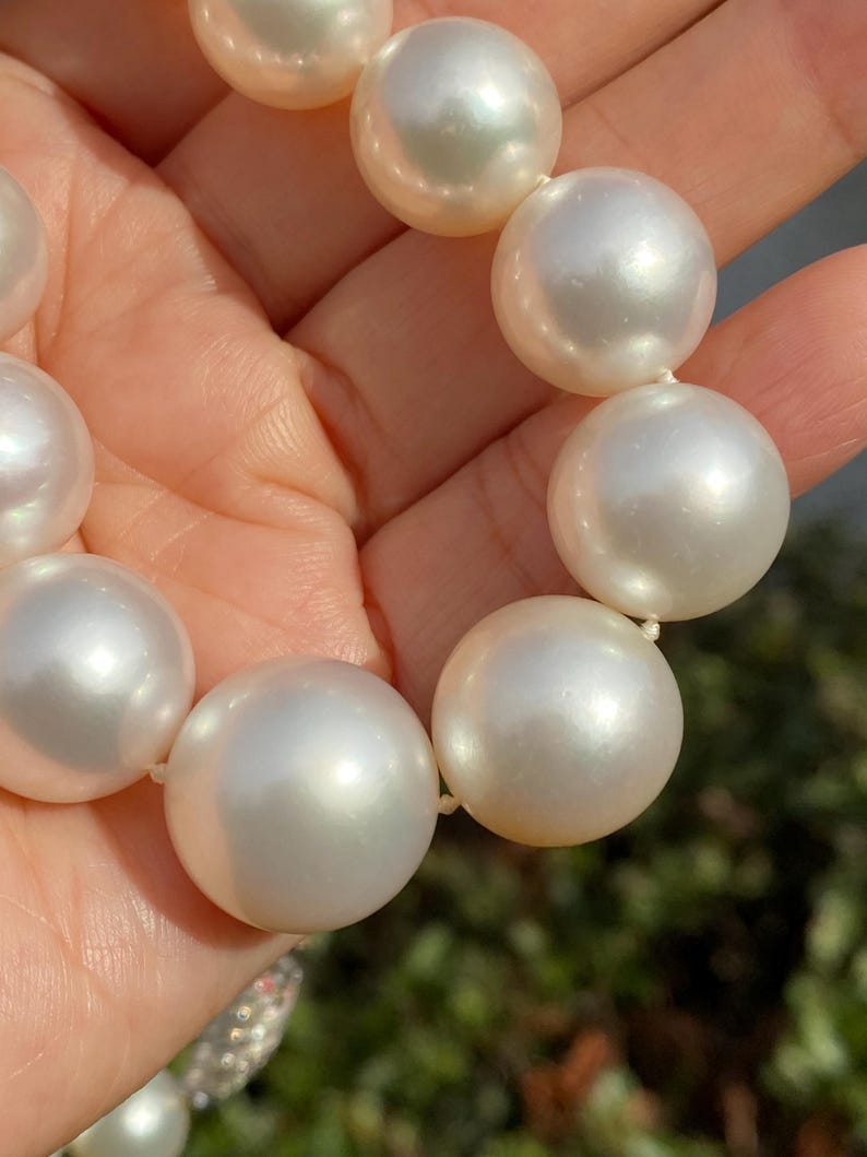 Pode incluir: Um colar de p&eacute;rolas brancas grandes em close-up. As p&eacute;rolas, de forma esf&eacute;rica, s&atilde;o de tamanho uniforme e t&ecirc;m um brilho perolado. Est&atilde;o enfiadas em um fio fino. O fundo desfocado sugere um ambiente externo.