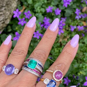 May include: A hand displays several gemstone rings. One ring features a large, oval, purple stone with two side stones. Another has a large emerald center stone, surrounded by diamonds and sapphires. A gold ring holds a pink cabochon, and another has a blue stone.