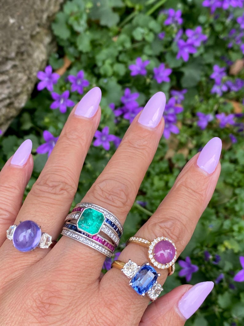 May include: A hand displays several gemstone rings. One ring features a large, oval, purple stone with two side stones. Another has a large emerald center stone, surrounded by diamonds and sapphires. A gold ring holds a pink cabochon, and another has a blue stone.