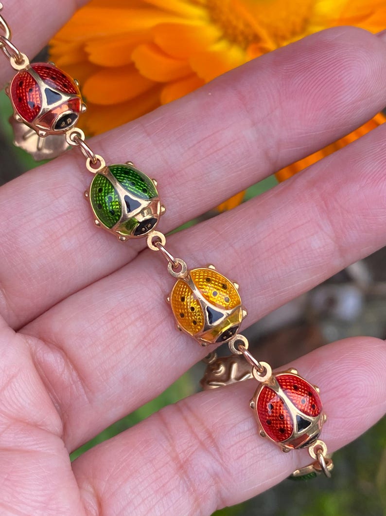May include: A gold chain bracelet with four ladybug charms. The ladybugs are red, green, yellow, and red with black spots.