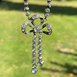 Op de afbeelding: Een zilverkleurige ketting met een bloemvormige hanger versierd met heldere edelstenen. De hanger heeft een centraal bloemmotief met bungelende edelsteenstrengen. De achtergrond is een wazig groen, wat duidt op een buitenomgeving.
