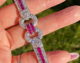 May include: A silver bracelet with a geometric design, featuring alternating sections of sparkling clear stones and vibrant red stones. The bracelet is held up against a blurred green background.