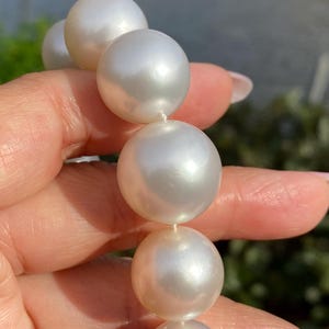 Pode incluir: Close-up de um colar de p&eacute;rolas brancas grandes e brilhantes. As p&eacute;rolas s&atilde;o redondas e de tamanho uniforme, enfiadas em um fio delicado. O fundo est&aacute; desfocado, destacando a superf&iacute;cie lisa e reflexiva das p&eacute;rolas. Joias.