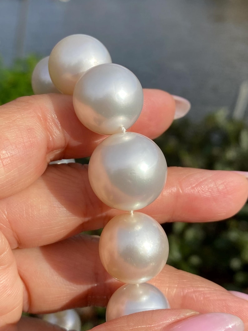 Pode incluir: Close-up de um colar de p&eacute;rolas brancas grandes e brilhantes. As p&eacute;rolas s&atilde;o redondas e de tamanho uniforme, enfiadas em um fio delicado. O fundo est&aacute; desfocado, destacando a superf&iacute;cie lisa e reflexiva das p&eacute;rolas. Joias.