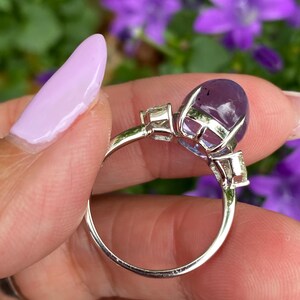May include: A silver ring featuring a large, oval-cut, purple gemstone as the centerpiece. Two smaller, rectangular, clear gemstones flank the main stone. The ring is held against a backdrop of purple flowers.