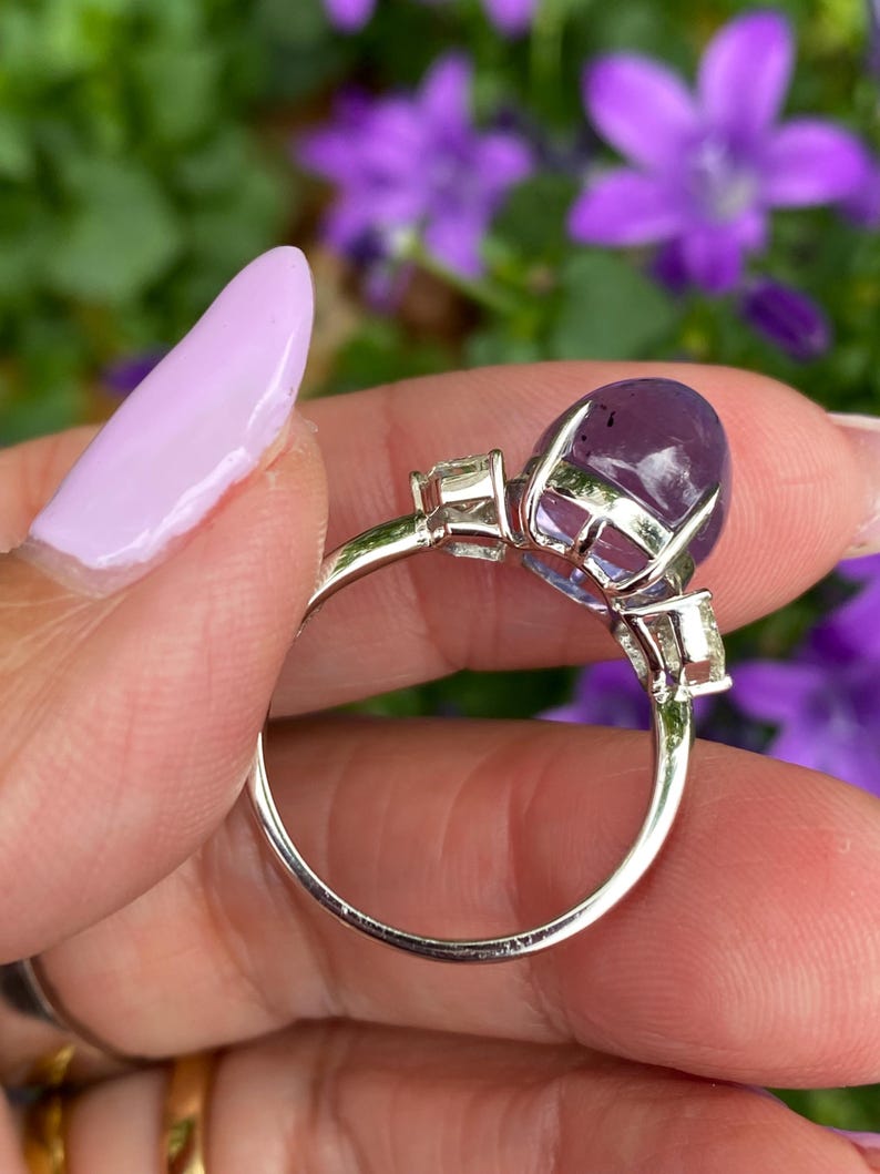 May include: A silver ring featuring a large, oval-cut, purple gemstone as the centerpiece. Two smaller, rectangular, clear gemstones flank the main stone. The ring is held against a backdrop of purple flowers.