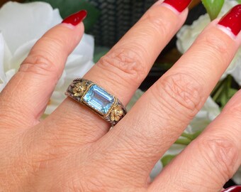 May include: A silver and gold ring with a rectangular, light blue gemstone. The ring features floral accents on the sides and is set on a hand with red nail polish.