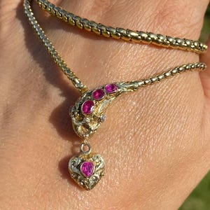 May include: A gold-toned necklace with a snake-like design. The necklace features a series of small, round links forming the chain. The snake's head is adorned with three pink gemstones, and a heart-shaped pendant with a pink gemstone hangs below.