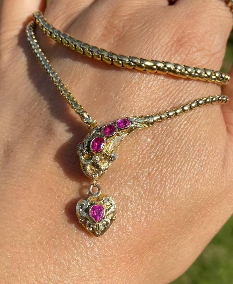 May include: A gold-toned necklace with a snake-like design. The necklace features a series of small, round links forming the chain. The snake's head is adorned with three pink gemstones, and a heart-shaped pendant with a pink gemstone hangs below.