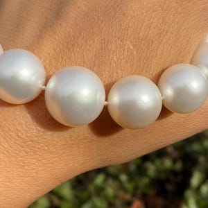 Pode incluir: Uma vista de perto de um colar com grandes p&eacute;rolas brancas e luminosas. As p&eacute;rolas esf&eacute;ricas s&atilde;o de tamanho uniforme e est&atilde;o enfiadas num fio branco fino. As p&eacute;rolas t&ecirc;m um brilho suave, que reflete a luz. O fundo est&aacute; desfocado, com toques de folhagem verde.