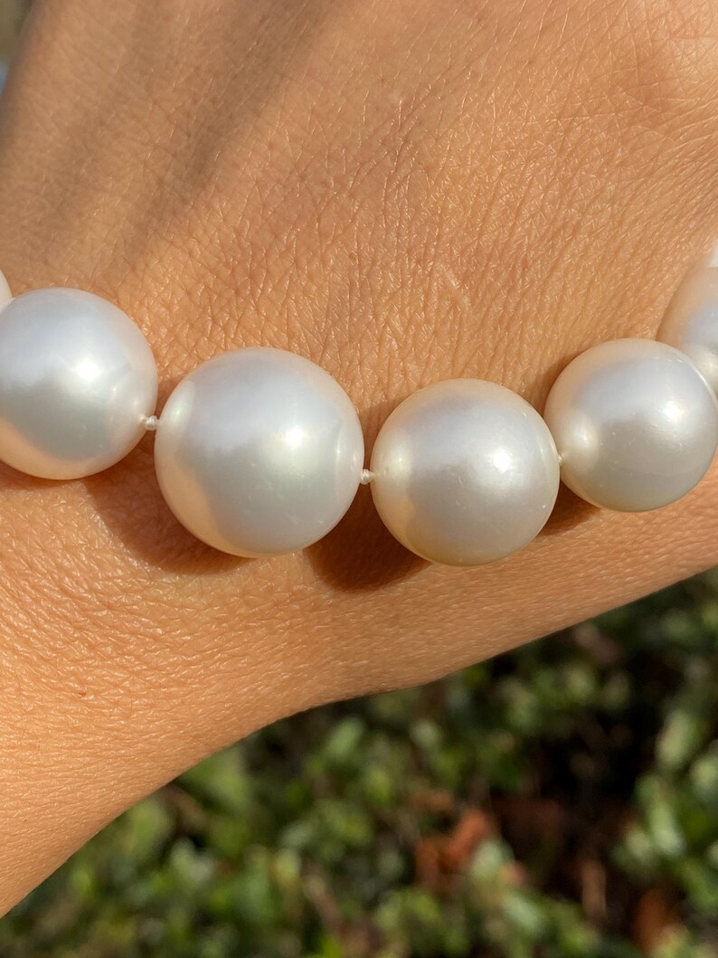 Pode incluir: Uma vista de perto de um colar com grandes p&eacute;rolas brancas e luminosas. As p&eacute;rolas esf&eacute;ricas s&atilde;o de tamanho uniforme e est&atilde;o enfiadas num fio branco fino. As p&eacute;rolas t&ecirc;m um brilho suave, que reflete a luz. O fundo est&aacute; desfocado, com toques de folhagem verde.