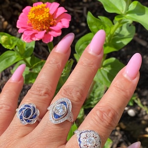 May include: Three silver rings featuring blue sapphires and clear stones. The first ring has a floral sapphire cluster, the second is an elongated marquise shape with a central stone, and the third is a scalloped design with a central stone.