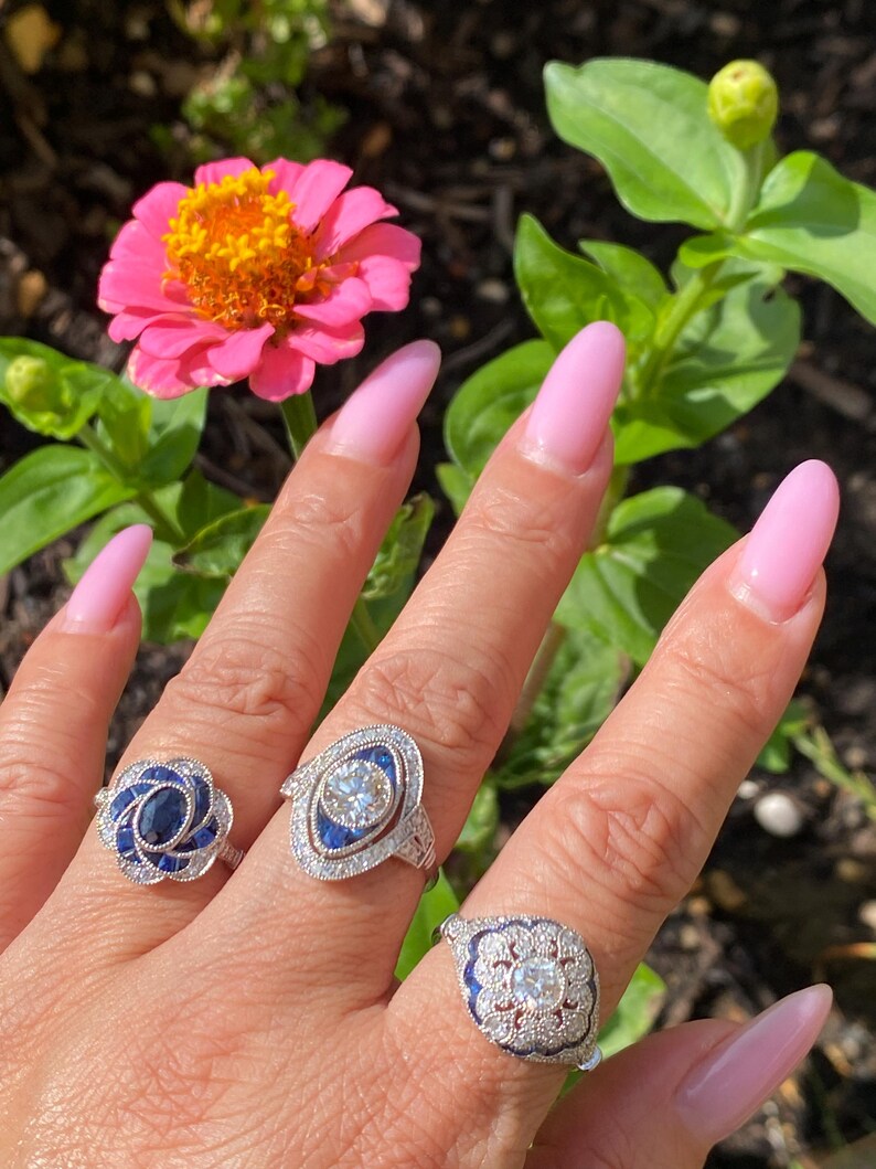 May include: Three silver rings featuring blue sapphires and clear stones. The first ring has a floral sapphire cluster, the second is an elongated marquise shape with a central stone, and the third is a scalloped design with a central stone.
