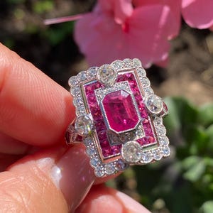 May include: A rectangular, Art Deco-style ring with a large, rectangular pink gemstone at the center, surrounded by smaller pink stones and clear, round accent stones. The ring has a silver-colored metal band.