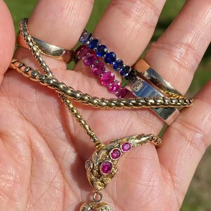 May include: A collection of gold jewelry, including a necklace with a ruby-studded snake pendant and a heart-shaped charm. Also visible are several rings, including a gold band and two gemstone eternity rings with blue and pink stones.