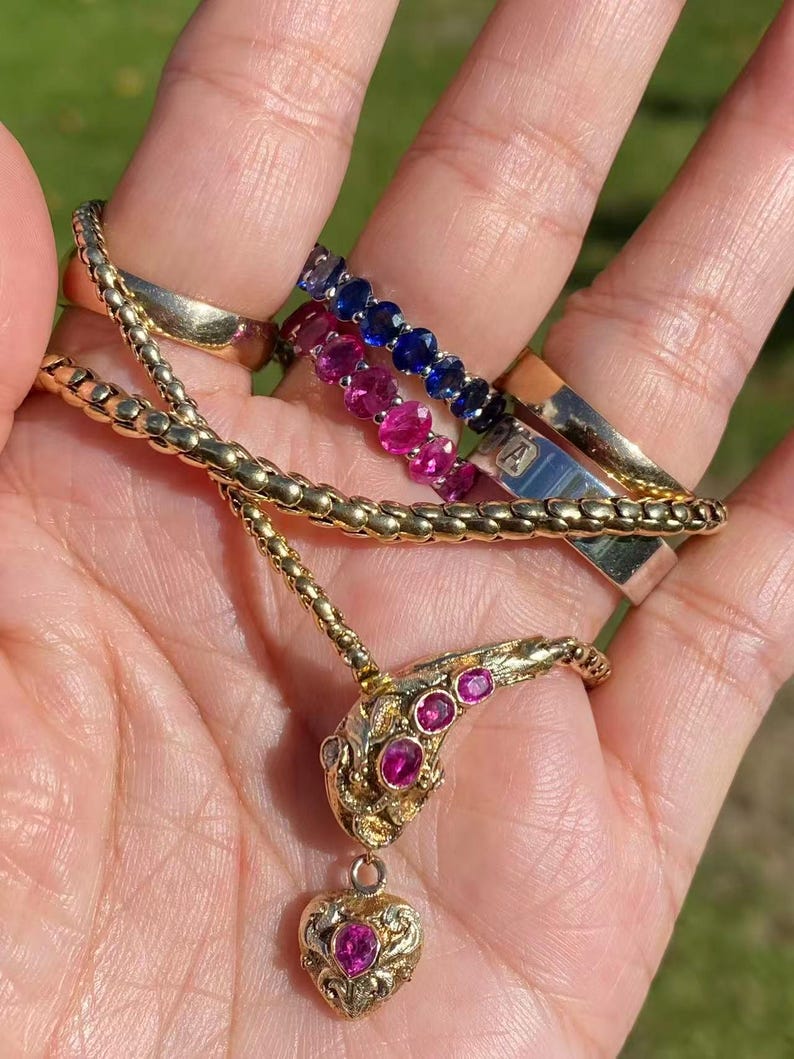 May include: A collection of gold jewelry, including a necklace with a ruby-studded snake pendant and a heart-shaped charm. Also visible are several rings, including a gold band and two gemstone eternity rings with blue and pink stones.