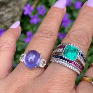 May include: Two rings are displayed on a finger. One ring features a large, oval, purple gemstone flanked by two clear, geometric stones. The other ring has a large, rectangular, emerald-green stone surrounded by smaller gems and bands of color.