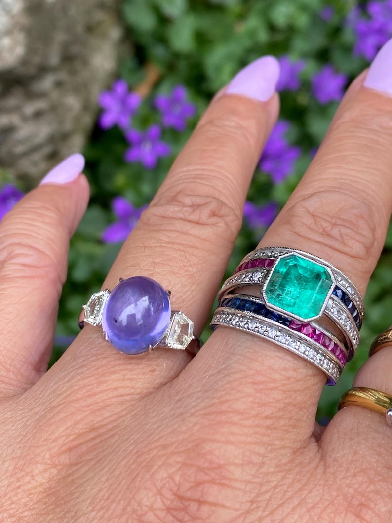 May include: Two rings are displayed on a finger. One ring features a large, oval, purple gemstone flanked by two clear, geometric stones. The other ring has a large, rectangular, emerald-green stone surrounded by smaller gems and bands of color.