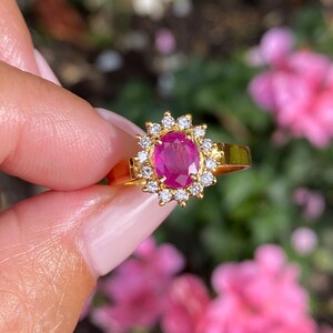 May include: A gold ring with an oval ruby centre stone, encircled by a halo of small diamonds. The ring's band is plain gold. The background shows pink flowers and green leaves, all softly blurred.