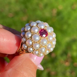 May include: A gold ring featuring a cluster of small, white pearls surrounding a central, round, red gemstone. The ring has a detailed, ornate design and is set against a blurred green background.