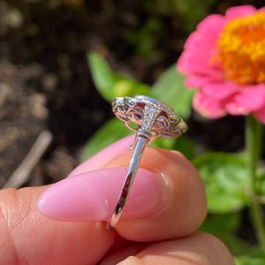 May include: A silver-toned ring featuring a central oval-shaped stone surrounded by a halo of smaller clear stones. The metal band displays intricate filigree detailing along the gallery and small stones set into the shoulders of the ring.